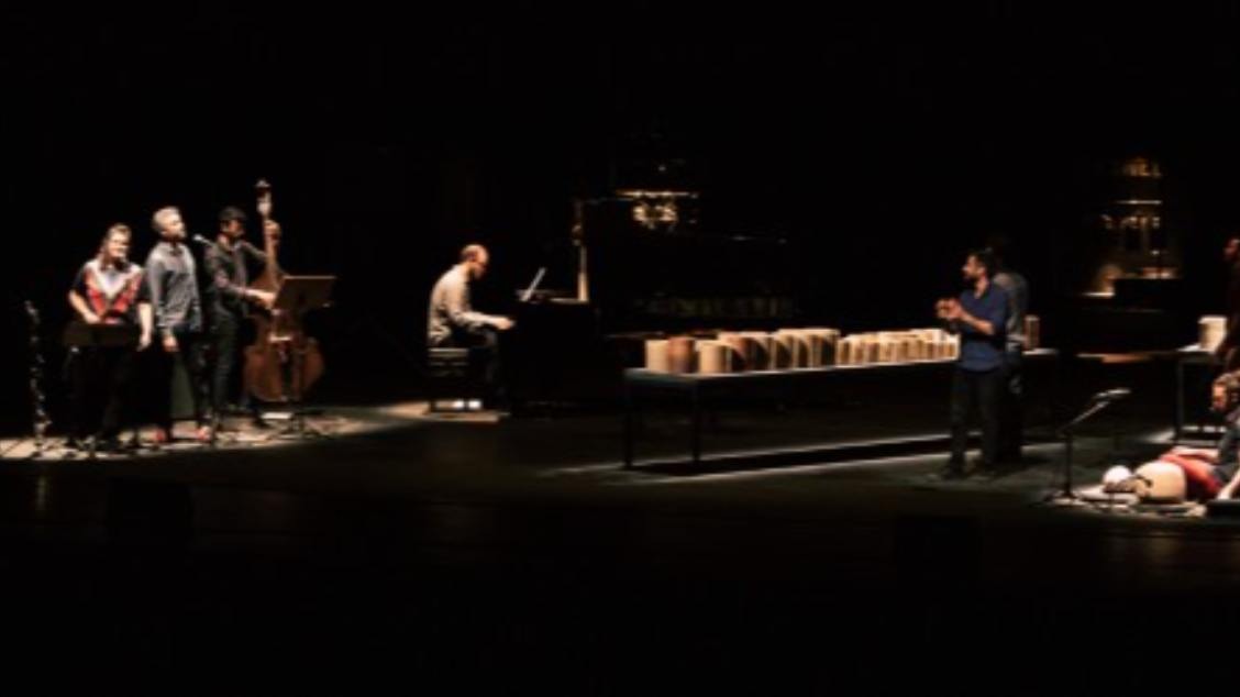 Cinco artistas no palco tocando instrumentos. Palco escuro com iluminação concetrada nos artistas.