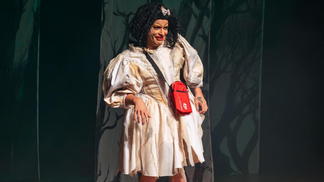 Suzy Brasil usando vestido branco com detalhes em amarelo, bolsa vermelha sobre os ombros e laço prata na cabeça. Imagem com fundo escuro.