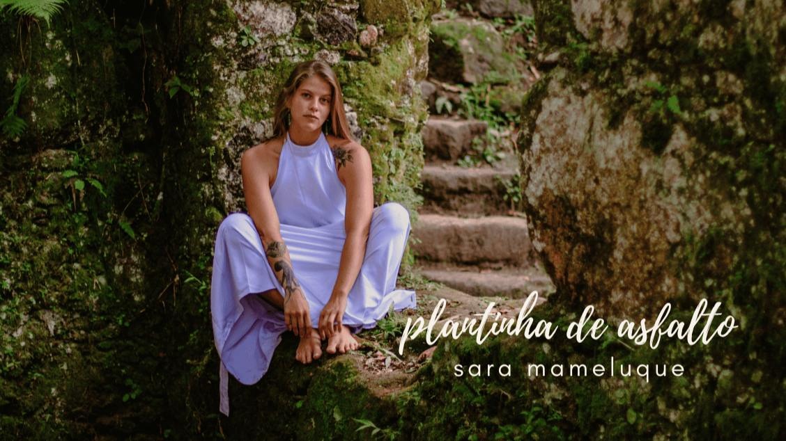Artista Sara Mameluque sentada em um chão de pedra coberto por vegetação rasteira. Ela usa vestido lilás e os pés descalõ, encara a câmera com semblante sério.