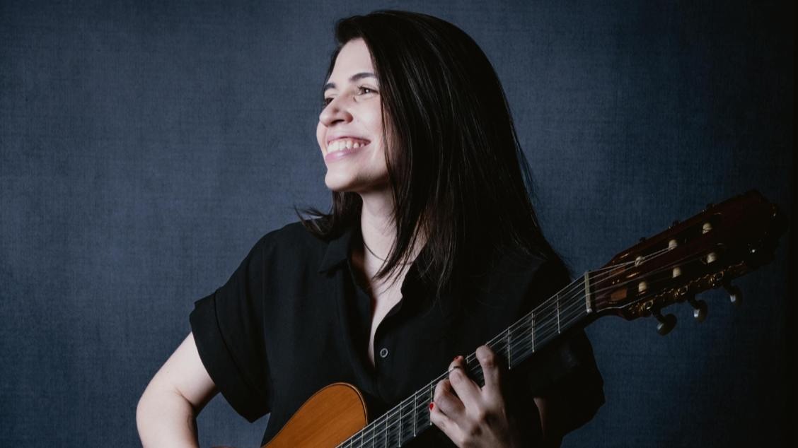 Foto: Divulgação/Centro Cultural Unimed-BH Minas Artista Marina Aquino vestindo blusa de botão preta e segurando um violão enquanto sorri. Imagem com fundo escuro.