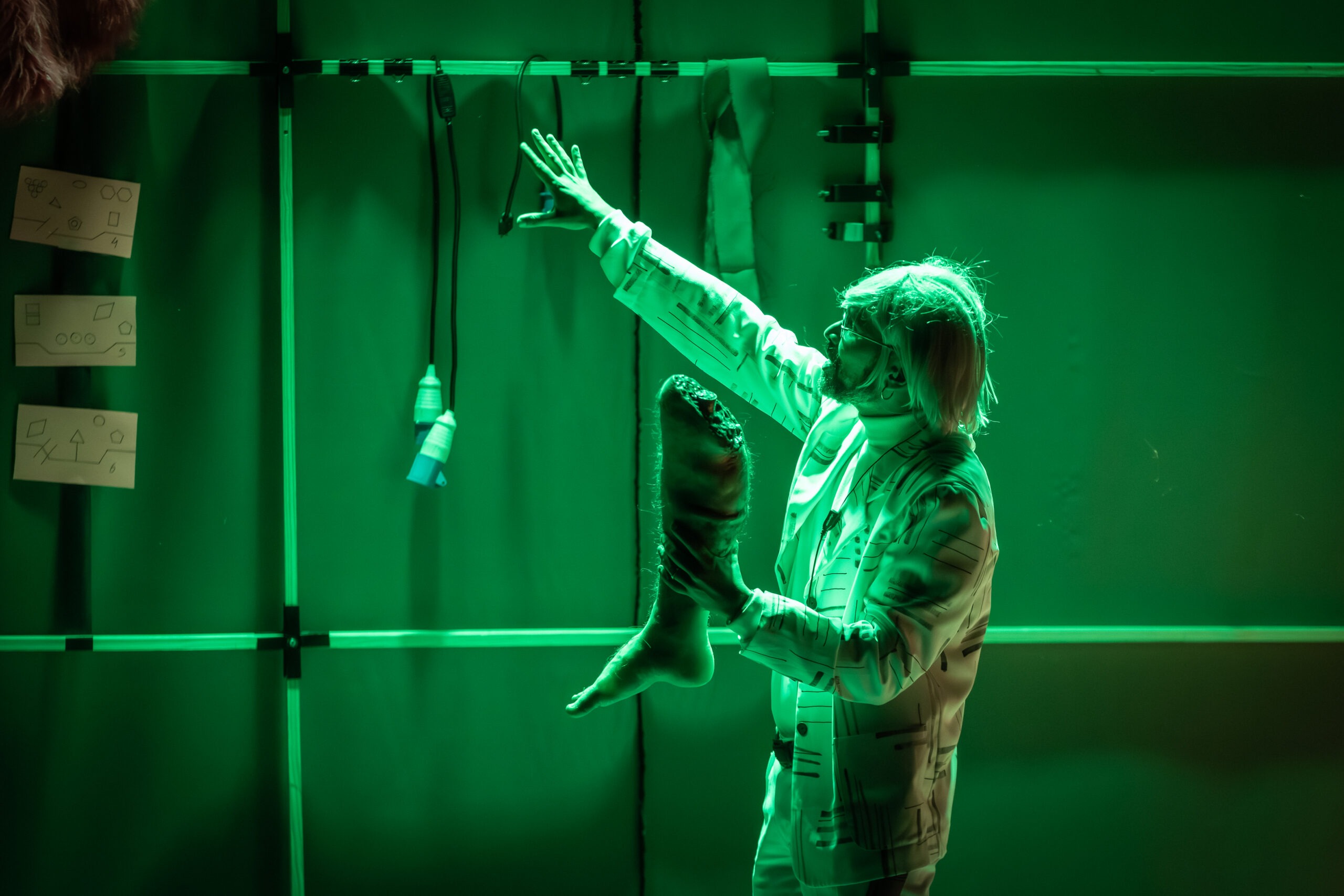 Homem segurando perna artificial humana e apontando para o alto com a outra mão. Veste roupas claras com manga longa e está sob luz verde.