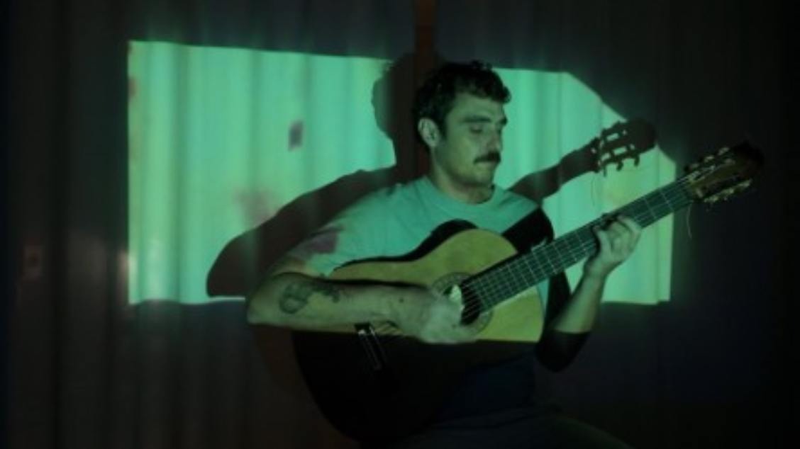 Artista Lucas Caram sentado tocando violão. Iluminação da foto direcionada ao artista.