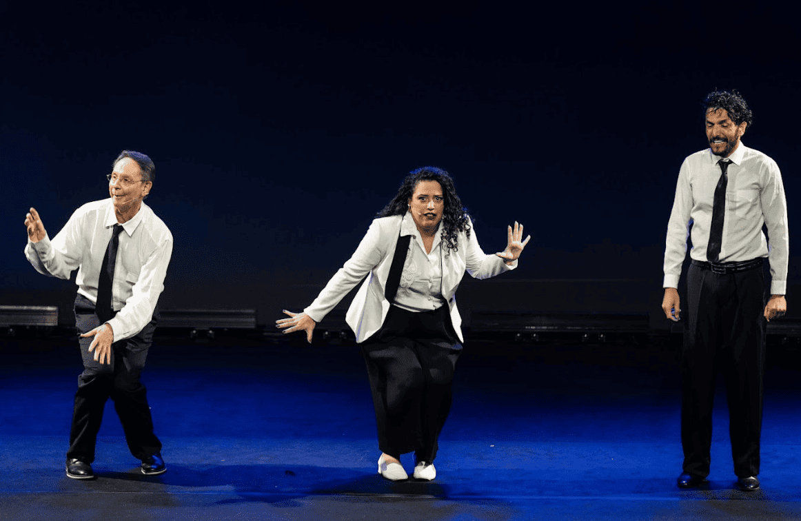 Três artistas — dois homens e uma mulher — realizam uma performance em um palco com fundo escuro e iluminação azulada. Todos vestem calças pretas e camisas brancas com gravatas. Eles exibem expressões dinâmicas e poses coreografadas, sugerindo um momento de teatro, dança ou improviso.