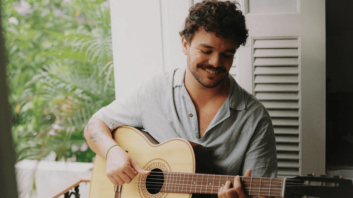A imagem, com tons suaves e naturais, mostra o músico sorrindo levemente enquanto olha para as cordas de um violão acústico. Ele veste uma camisa listrada de manga curta e está posicionado em frente a uma janela com venezianas brancas abertas. Ao fundo, à esquerda, é possível ver a folhagem verde de palmeiras, criando uma ambientação tranquila e iluminada pela luz do dia.