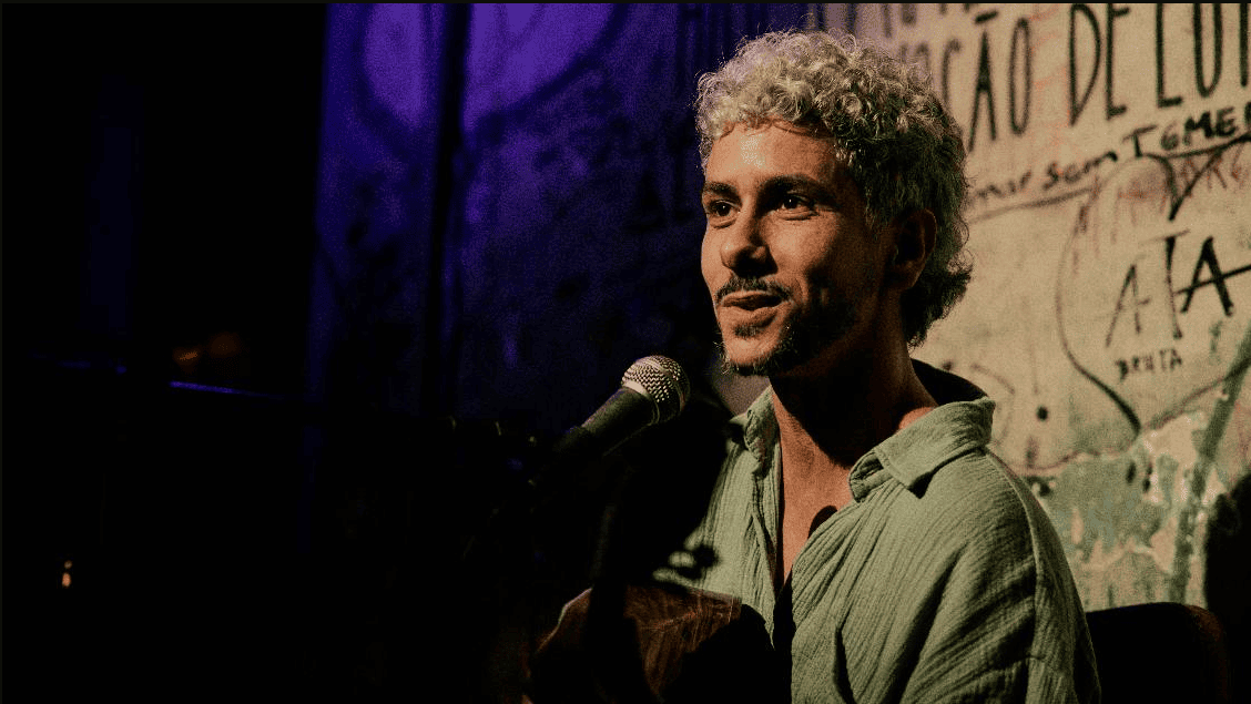 A imagem mostra o artista de perfil parcial, sorrindo suavemente enquanto canta em um microfone e toca violão. Ele tem cabelos curtos, cacheados e descoloridos, e veste uma camisa de tecido leve na cor verde-água. O fundo é escuro, com uma iluminação dramática que destaca tons de roxo à esquerda e uma parede com pichações e frases artísticas à direita, criando uma estética urbana e acolhedora de show ao vivo.