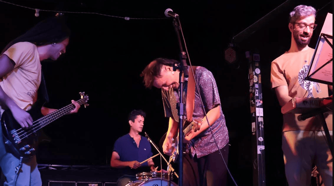 A imagem captura quatro músicos em um palco com iluminação focada e fundo escuro. No centro, o guitarrista — com uma camisa brilhante prateada — aparece inclinado sobre seu instrumento em um momento de entrega. À esquerda, um baixista sorri enquanto toca; ao fundo, o baterista mantém o ritmo concentrado; e à direita, o tecladista acompanha a performance com uma partitura à frente. O clima é de descontração e sintonia entre os artistas durante o show.