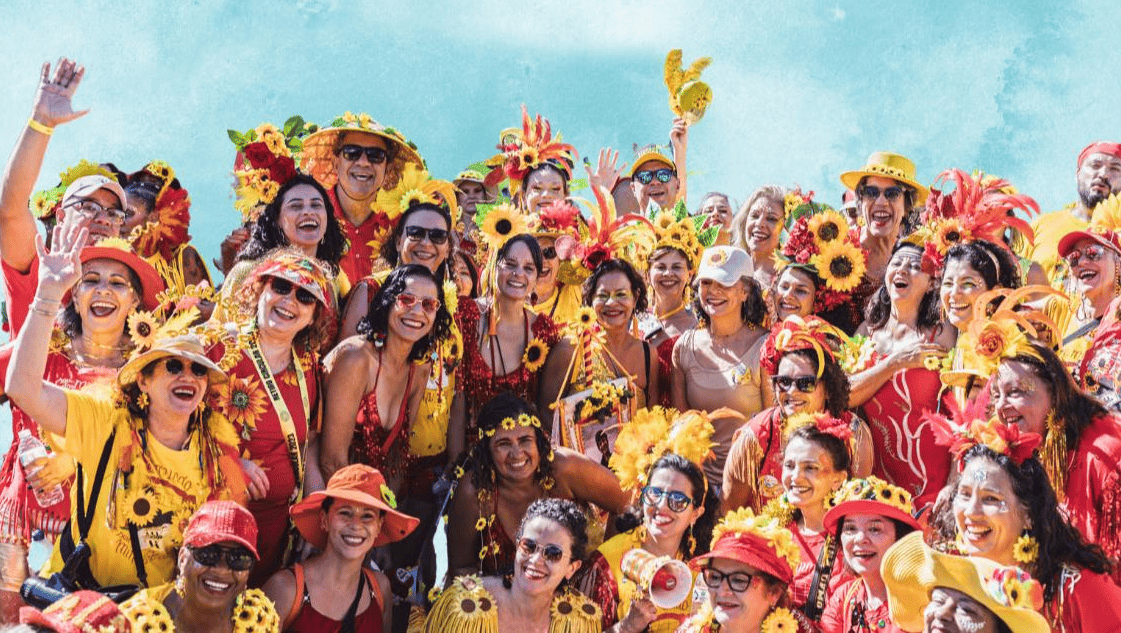A imagem mostra um grande grupo de foliões sorridentes, a maioria mulheres, celebrando o Carnaval. Eles vestem fantasias vibrantes nas cores vermelho e amarelo, com destaque para diversos adereços de girassóis em chapéus, tiaras e roupas. A atmosfera é de alegria e união, com várias pessoas acenando para a câmera sob um céu azul claro ao fundo. É uma representação típica da energia festiva dos blocos de rua mineiros.