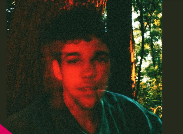Fotografia em estilo granulado de um jovem de pele clara e cabelos escuros, com um efeito de dupla exposição que deixa seu rosto avermelhado. Ele está em uma floresta, com troncos de árvores e folhagens ao fundo iluminados pela luz do sol.