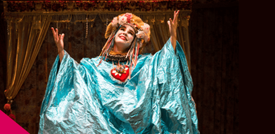 Imagem horizontal mostra uma atriz no palco, do busto para cima, caracterizada com um figurino teatral exuberante. Ela é uma mulher branca, com o rosto pintado de branco e bochechas rosadas, que sorri olhando para o alto com os braços abertos e as mãos erguidas. Veste uma túnica larga e brilhante em tom azul-celeste, um colar com um grande coração vermelho decorado no centro do peito e um adereço de cabeça dourado adornado com flores e fitas coloridas pendentes.