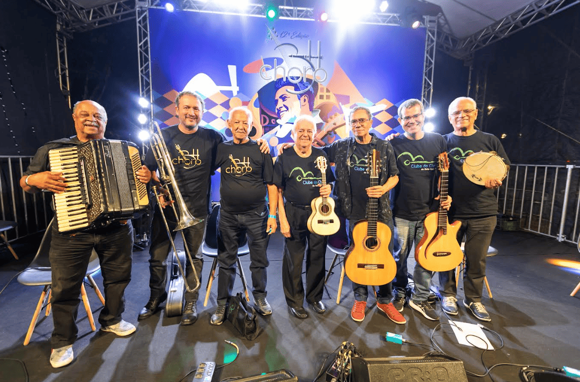 Fotografia colorida de sete músicos homens, adultos e idosos, posando sorridentes lado a lado no palco iluminado da 12ª Edição do BH Choro. Eles vestem calças e camisetas pretas do evento e seguram, da esquerda para a direita: um acordeon, um trombone de vara, um cavaquinho, dois violões e um pandeiro (o terceiro integrante está sem instrumento). Ao fundo, destaca-se um grande painel azul com a ilustração estilizada do músico Pixinguinha tocando flauta.