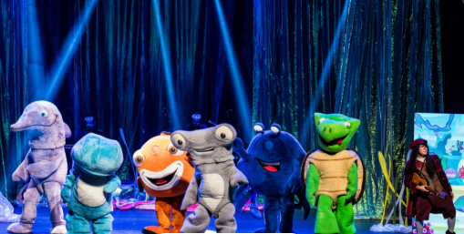Foto de uma apresentação teatral infantil no palco. Ao centro, seis atores vestem fantasias grandes e coloridas de animais marinhos: um golfinho cinza, um peixe azul, um peixe-palhaço laranja, um tubarão-martelo cinza, um caranguejo azul-escuro e uma tartaruga verde. No canto direito, uma atriz caracterizada como pirata segura um instrumento musical. O cenário ao fundo tem cortinas brilhantes esverdeadas iluminadas por feixes de luz azul.