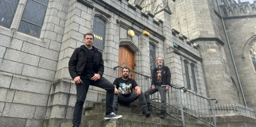 Fotografia colorida tirada de baixo para cima, mostrando três homens com roupas escuras e estilo casual/rocker posando nas escadarias de uma antiga construção de pedras cinzas, que lembra uma igreja gótica. Um homem está de pé à esquerda com a mão na cintura, outro está sentado ao centro dos degraus, e o terceiro está de pé à direita, perto de um corrimão de metal. Ao fundo, destacam-se uma porta de madeira em arco e janelas altas com vitrais.