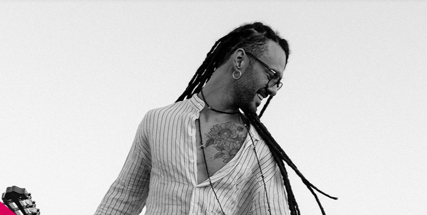 Fotografia em preto e branco com fundo claro. Um homem jovem, com pele negra clara, sorri de perfil olhando para baixo e para a direita. Ele tem cabelos longos com dreadlocks que caem pelas costas e ombros, usa óculos de grau, brinco de argola e um colar discreto. Veste uma camisa clara com listras verticais finas, que está entreaberta, revelando uma tatuagem de girassóis no peito. No canto inferior esquerdo, aparece uma pequena parte do braço de um violão.