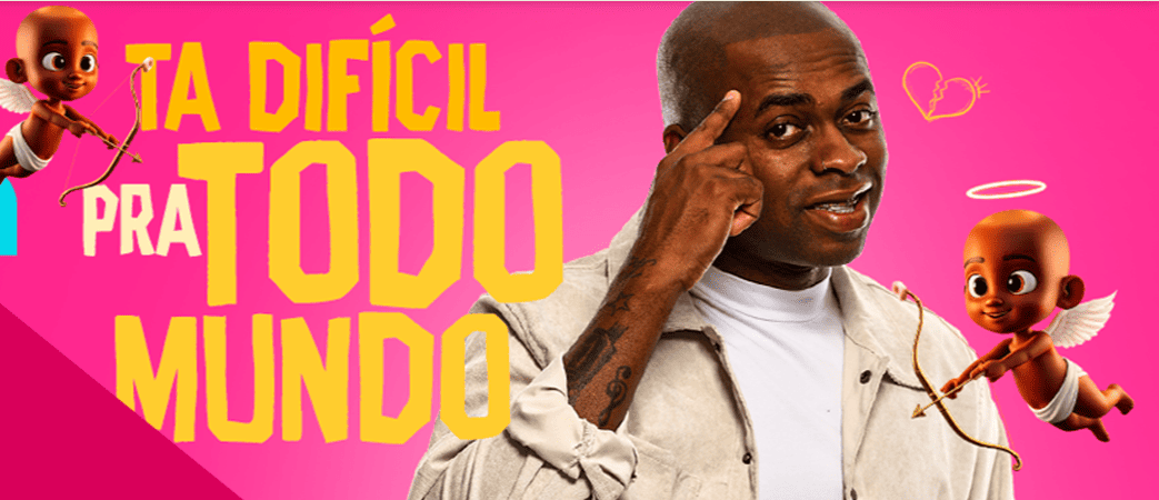 Imagem promocional com fundo rosa vibrante onde se destaca, à esquerda, o texto "TÁ DIFÍCIL PRA TODO MUNDO" escrito em letras amarelas e brancas. No centro, um homem negro sorri apontando o dedo indicador para a própria cabeça. Ele está cercado por dois pequenos cupidos negros de asas brancas e fralda segurando arco e flecha, além de um desenho de coração partido na parte superior.