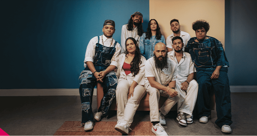 Foto em estúdio de um grupo de oito pessoas vestindo roupas jeans e brancas, posando juntas diante de uma parede dividida entre as cores azul e bege.