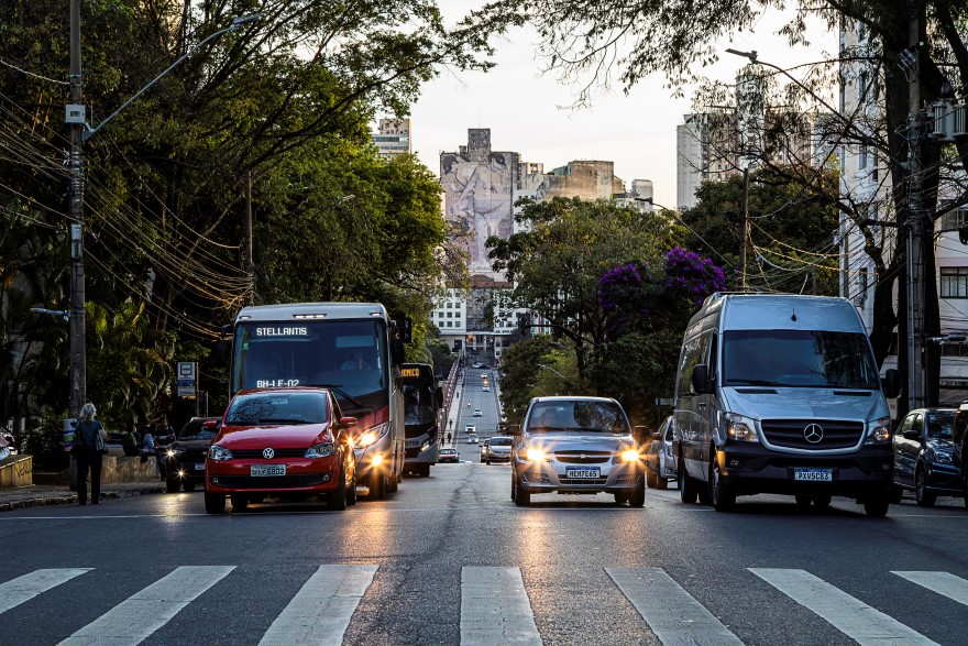 Pablo Gomide/Quarteto Filmes uma avenida movimentada com carros e ônibus em um final de tarde. Na rua, no sentido do observador, há uma van prateada à direita e um ônibus urbano à esquerda. Ao fundo, no horizonte, é possível ver um edifício com um grande mural com duas mulheres desenhadas, em meio à paisagem urbana de prédios e árvores.