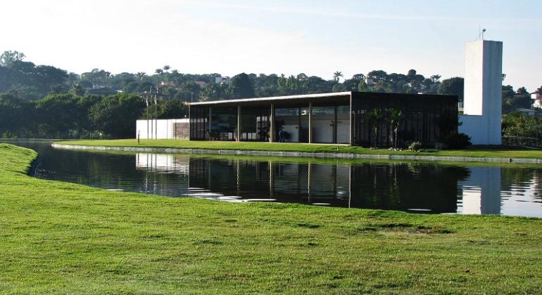 Edifício moderno e retangular no Parque Ecológico da Pampulha. O prédio, de cor escura, tem grandes janelas de vidro e uma torre branca ao lado. Ele está à beira de um lago que reflete sua imagem. O primeiro plano é uma vasta área de grama verde que se estende até a água.