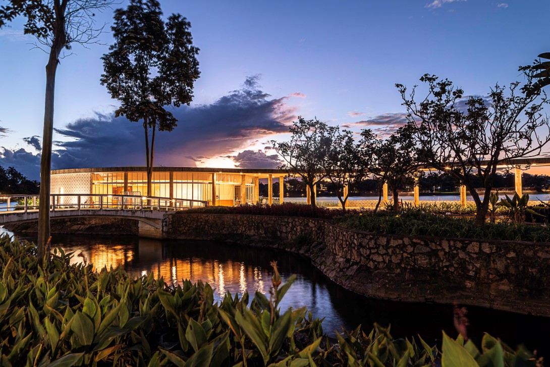 Casa do Baile ao anoitecer. É uma construção redonda e moderna, iluminada por dentro, que se estende sobre a água, conectada por uma ponte curva. O céu está em tons de azul, roxo e laranja do pôr do sol. A vegetação verde no primeiro plano reflete na água escura.