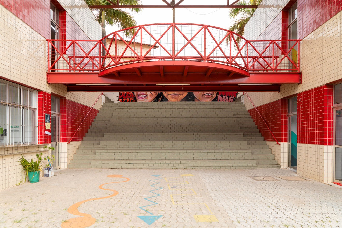Entrada interna com escadaria larga de blocos cinzas que leva ao andar superior. Acima, há uma passarela vermelha metálica com grades. No fundo, parte de um mural colorido com rostos é visível.