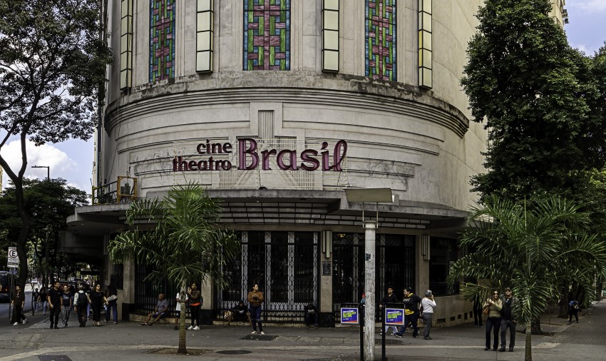 Edifício Cine Theatro Brasil, uma construção histórica em Belo Horizonte. No centro da foto, a fachada curva do prédio é visível. A entrada, ao nível da rua, possui grandes portas de vidro e metal. Acima da entrada, a fachada apresenta a inscrição "cine theatro Brasil" com letras de cor rosa, e detalhes arquitetônicos em relevo. Na parte superior da construção, grandes vitrais retangulares, com padrões coloridos em tons de verde, rosa e azul, se destacam. Em frente ao prédio, há palmeiras e pessoas.