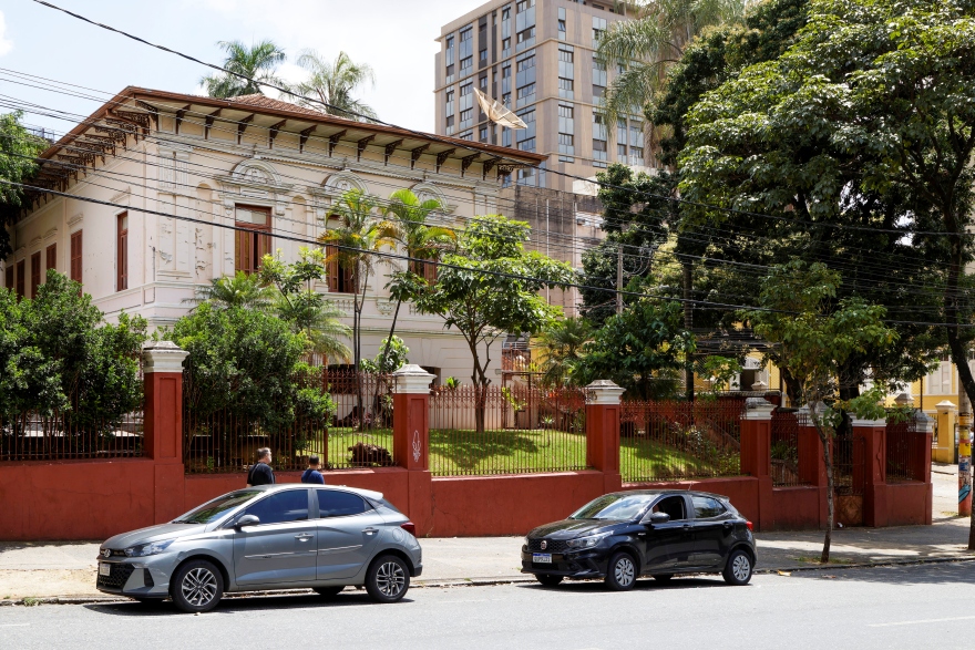 Imagem mostra a fachada de um casarão antigo de dois andares, pintado de branco com detalhes em vermelho, cercado por muro e grades também vermelhas. Em frente ao prédio há árvores, arbustos e duas pessoas caminhando na calçada. Dois carros estão estacionados na rua, um cinza e um preto. Ao fundo aparece um prédio moderno de vários andares e outras árvores grandes. Fios elétricos atravessam a parte superior da cena.