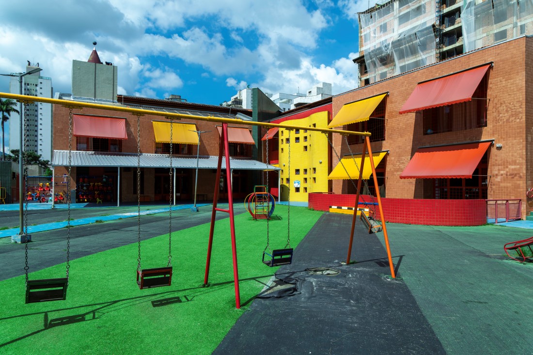 Pátio externo com um balanço de estrutura vermelha e laranja em primeiro plano, sobre uma área de grama sintética. Ao fundo, a fachada da escola em tijolo aparente, com toldos vermelhos e amarelos, e um bloco central amarelo com pequenas janelas quadradas. Prédios altos cercam o local.