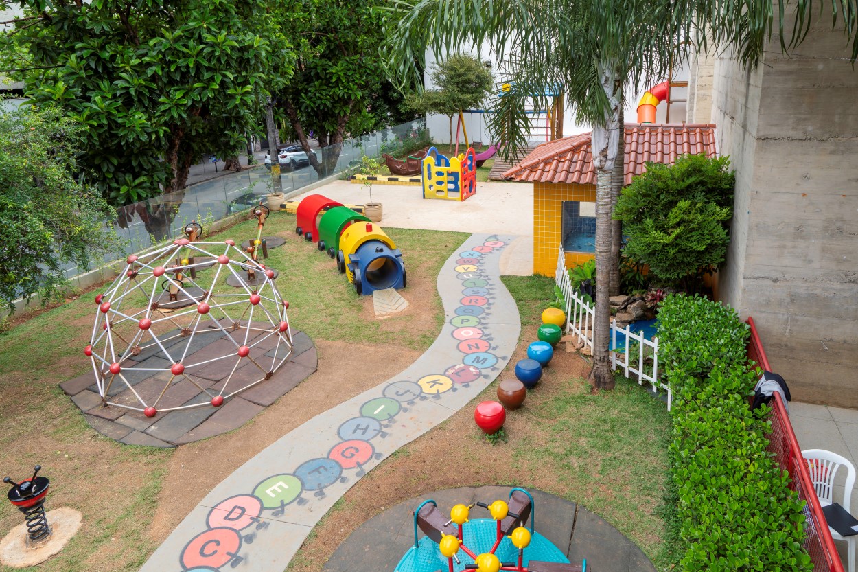 Vista de cima de um grande parquinho gramado com diversos brinquedos. Um caminho de cimento com letras coloridas do alfabeto serpenteia pelo jardim. Há um túnel em formato de centopeia, um gira-gira, um domo de escalada e uma casinha de brinquedo amarela com telhado de cerâmica.