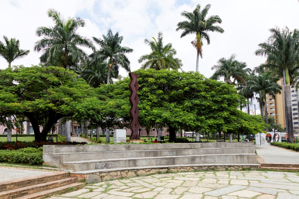 Em primeiro plano, um palco com três degraus feitos de cimento, com o chão à frente coberto por lajes de pedra irregular. Acima, no centro, uma escultura escura, de formato orgânico e retorcido, emerge entre duas árvores de copa densa e verde. Ao fundo, muitas palmeiras altas e edifícios