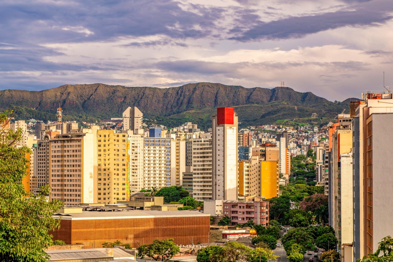 Vista panorâmica da cidade com prédios altos no centro e montanha ao fundo. No primeiro plano, árvores verdes parcialmente encobrem parte da cena.