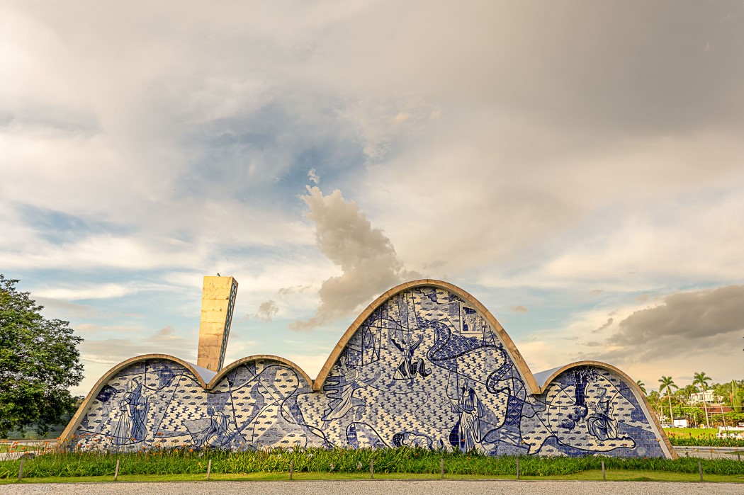 Uma igreja com estrutura arquitetônica de forma ondulada, coberta por azulejos azuis e brancos em padrão de mosaico, representando cenas históricas e figuras humanas. Ao fundo, um céu nublado com tons de cinza e azul claro. À esquerda, uma torre retangular de concreto. Em primeiro plano, grama verde e uma calçada. Palmeiras e árvores ao longe.