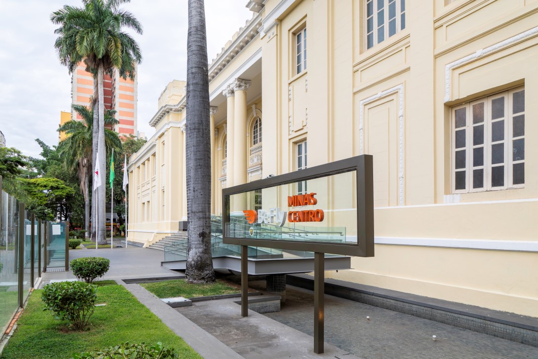 O BEFLY Minas Centro, em BH, exibe sua fachada creme clássica com janelas brancas e colunas. No primeiro plano, um totem moderno de vidro e metal, com o nome "BEFLY MINAS CENTRO" em laranja, destaca a entrada. Palmerias altas e coqueiros emolduram a área, com gramados verdes e um edifício ao fundo.