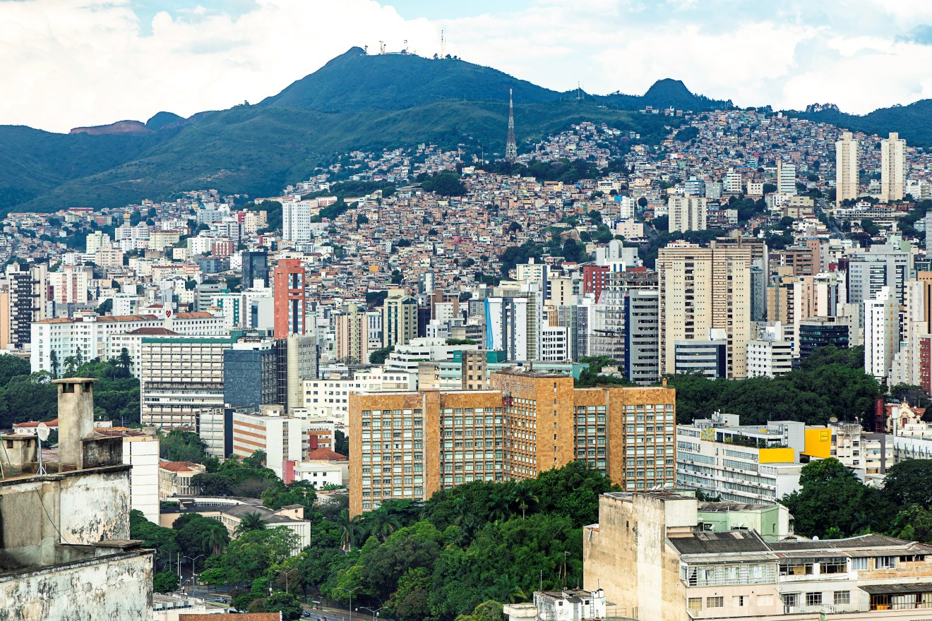 Vista panorâmica da cidade de Belo Horizonte, com prédios altos, casas em morro e a Serra do Curral ao fundo. Em primeiro plano, edifício de fachada bege e marrom. Céu parcialmente nublado acima.