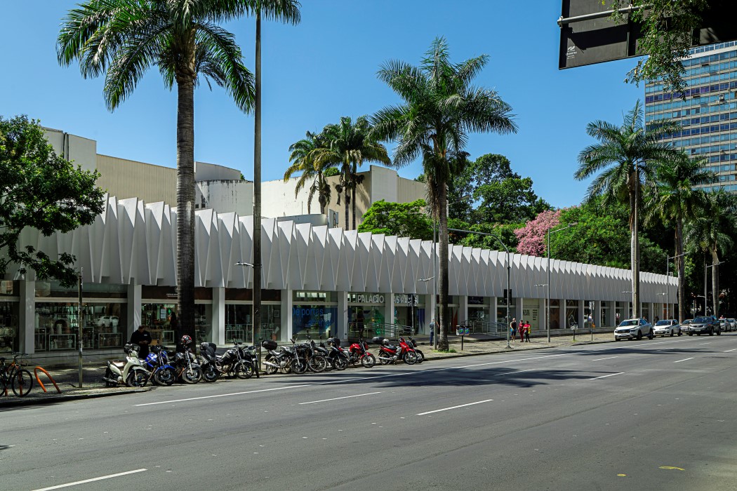 Fachada frontal do Palácio das Artes. O prédio tem uma estrutura longa e horizontal, com grandes janelas de vidro e uma parede branca em ziguezague no andar de cima. Uma rua larga está em primeiro plano, com motos e carros estacionados na calçada, que é ladeada por palmeiras. O céu está azul e sem nuvens.