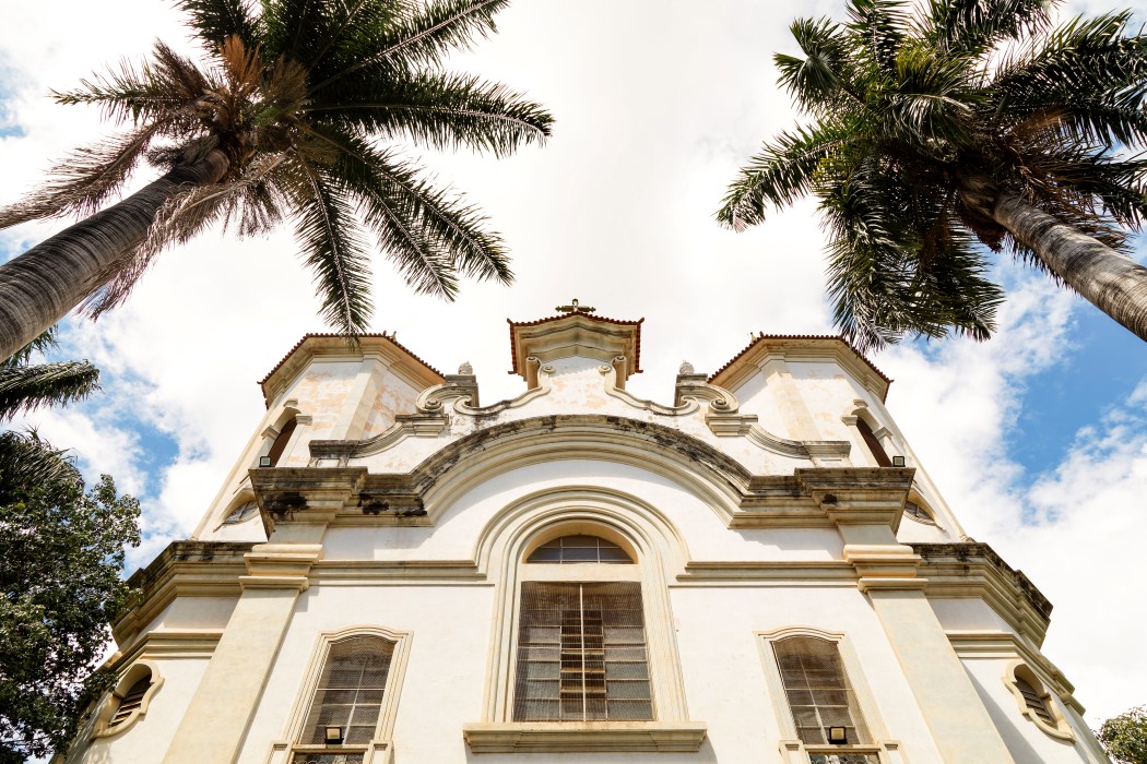 de baixo pra cima da Paróquia Santa Teresa e Santa Teresinha. A igreja, de cor clara, tem um estilo clássico com janelas em arco. No primeiro plano, duas grandes palmeiras com troncos grossos enquadram a igreja. O céu está azul com grandes nuvens brancas e claras.