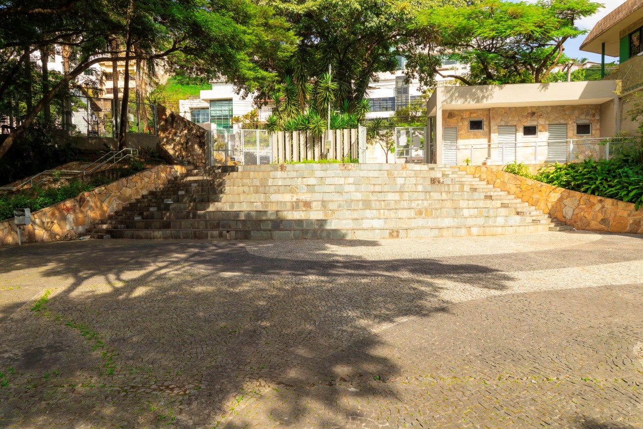 : Escadaria larga de pedra ocupa o centro da imagem, composta por degraus retos e lisos. Nas laterais, muros inclinados de pedra acompanham a subida. No alto, há um portão metálico fechado, atrás do qual se vê vegetação e parte de um prédio. À direita, um prédio baixo com portas e janelas dá vista para o pátio.