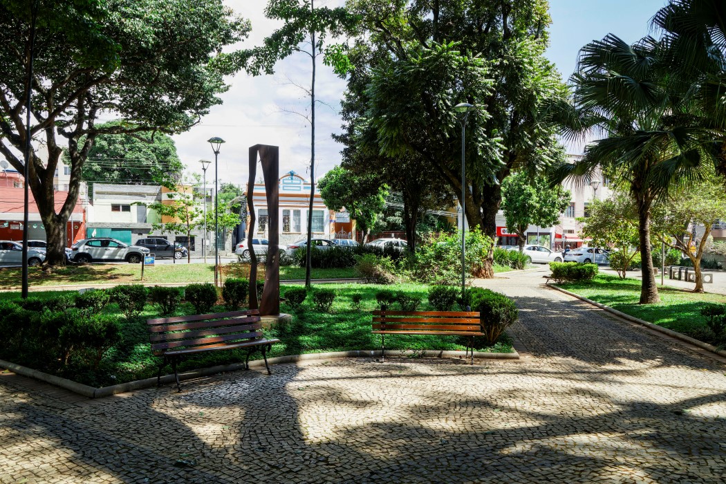 Uma praça com piso de pedras portuguesas e canteiros de grama e arbustos. Dois bancos de madeira com pés de ferro estão posicionados de frente, um à esquerda e outro à direita. Ao centro, entre árvores e postes de iluminação, há uma escultura de metal vertical e enferrujado. Ao fundo, veem-se ruas e casas.