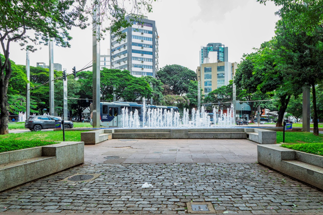 Praça com chafariz central, jatos de água brancos saem de um tanque retangular cercado por bancos de concreto. Ao fundo, prédios altos, ônibus e árvores verdes. Piso de pedra e concreto ocupa o primeiro plano.