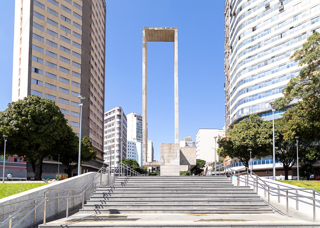 Vista a partir da base de uma longa escadaria de granito que leva à praça. O grande monumento de concreto em formato de portal está centralizado no topo da escada, enquadrado por dois altos edifícios nas laterais. O céu está limpo e com uma tonalidade de azul claro e uniforme.