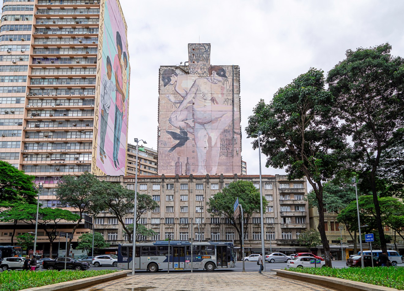 Vista frontal da praça, com canteiro de plantas verdes e árvores altas. Ao fundo, grande mural em prédio mostrando figuras humanas. À esquerda, prédio com fachada clara e outro com mural colorido. Ônibus e carros passam na avenida em frente.