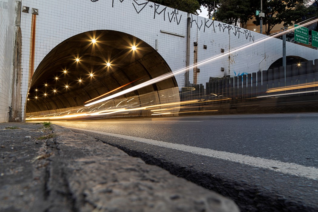 Vista em ângulo baixo da entrada do túnel à noite. A entrada é um arco revestido por azulejos brancos pichados. Dentro do túnel, luzes amarelas criam um efeito de estrela. Rastros de luz de veículos, em tons de branco e laranja, entram e saem do túnel, cruzando a imagem na horizontal.