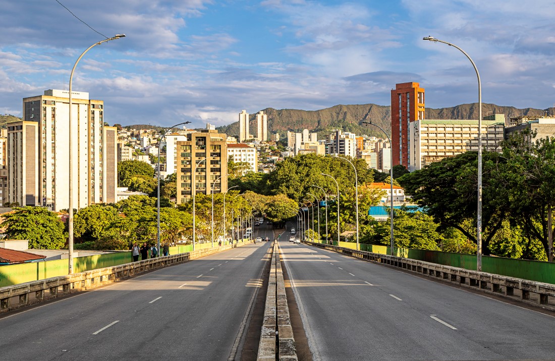 Vista a partir do centro de um viaduto com múltiplas pistas de asfalto. A via, vazia, se estende em perspectiva em direção a uma área urbana com prédios e muitas árvores. Ao fundo, uma cadeia de montanhas sob um céu azul. Postes de iluminação curvos ladeiam o viaduto.