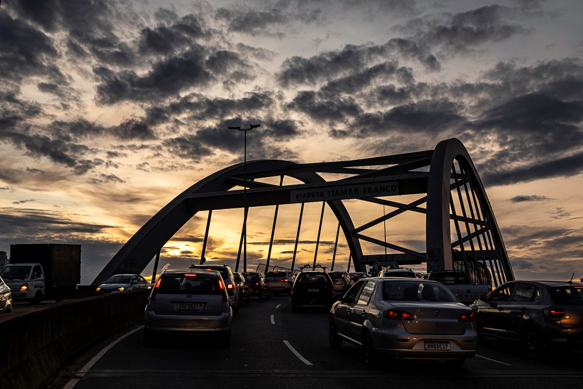 Trânsito intenso sobre o viaduto, com vários carros vistos por trás. A estrutura em arco do viaduto, com a inscrição "VIADUTO ITAMAR FRANCO", está centralizada. O céu está preenchido por nuvens escuras e dramáticas, com a luz alaranjada do crepúsculo surgindo no horizonte sob o arco.