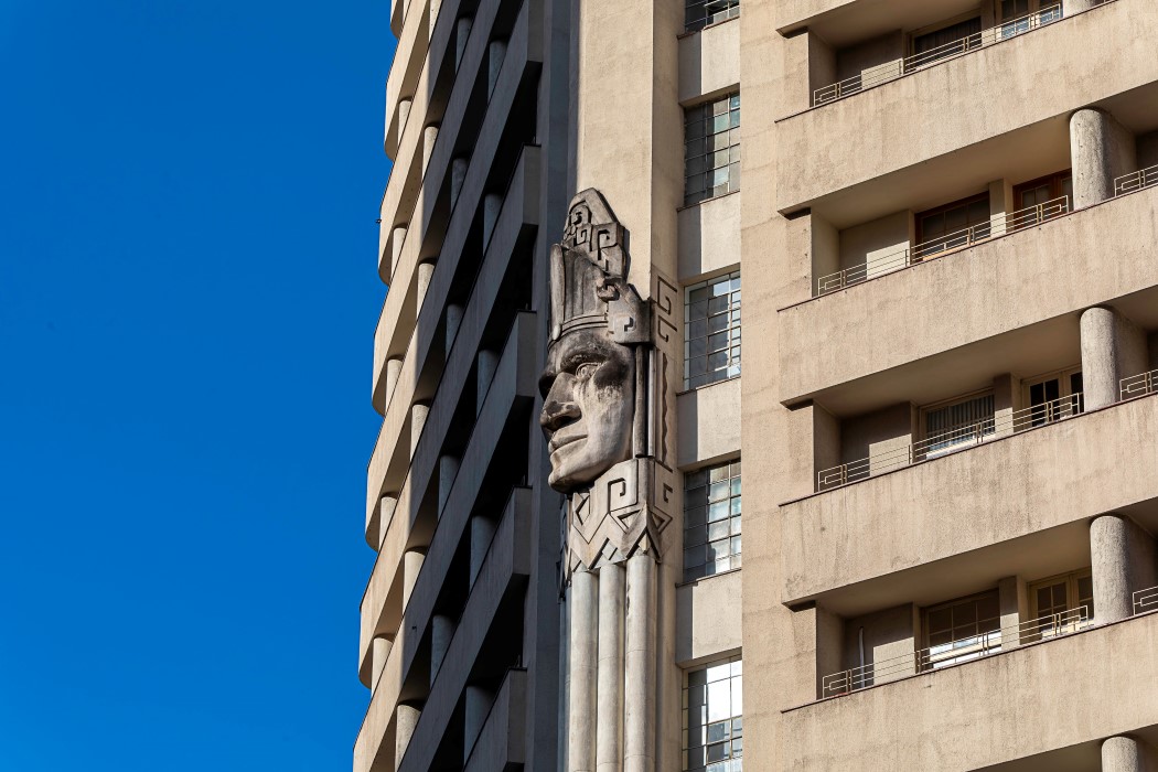 Um close na lateral do Edifício Acaiaca, mostrando um detalhe arquitetônico em relevo. No centro, uma escultura gigante de uma cabeça indígena esculpida em pedra ou concreto se destaca. A cabeça, com um cocar, está em uma parede estreita entre duas seções diferentes do prédio.