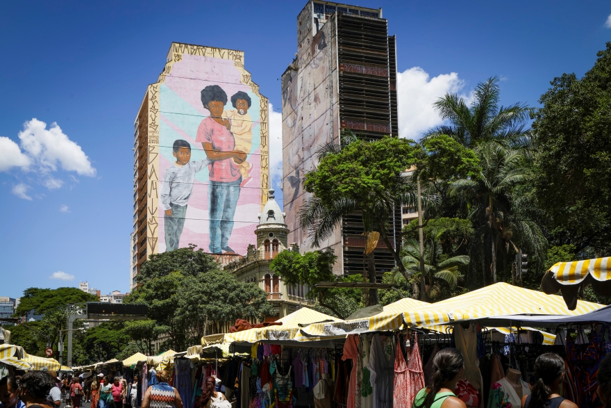 Pablo Gomide/Quarteto Filmes Imagem de uma feira ao ar livre com barracas de toldos listrados amarelo e branco. Pessoas circulam entre roupas expostas para a venda. Ao fundo, um prédio alto exibe um grande mural com a pintura de uma mulher negra carregando um bebê e duas crianças ao lado. Árvores cercam a cena.