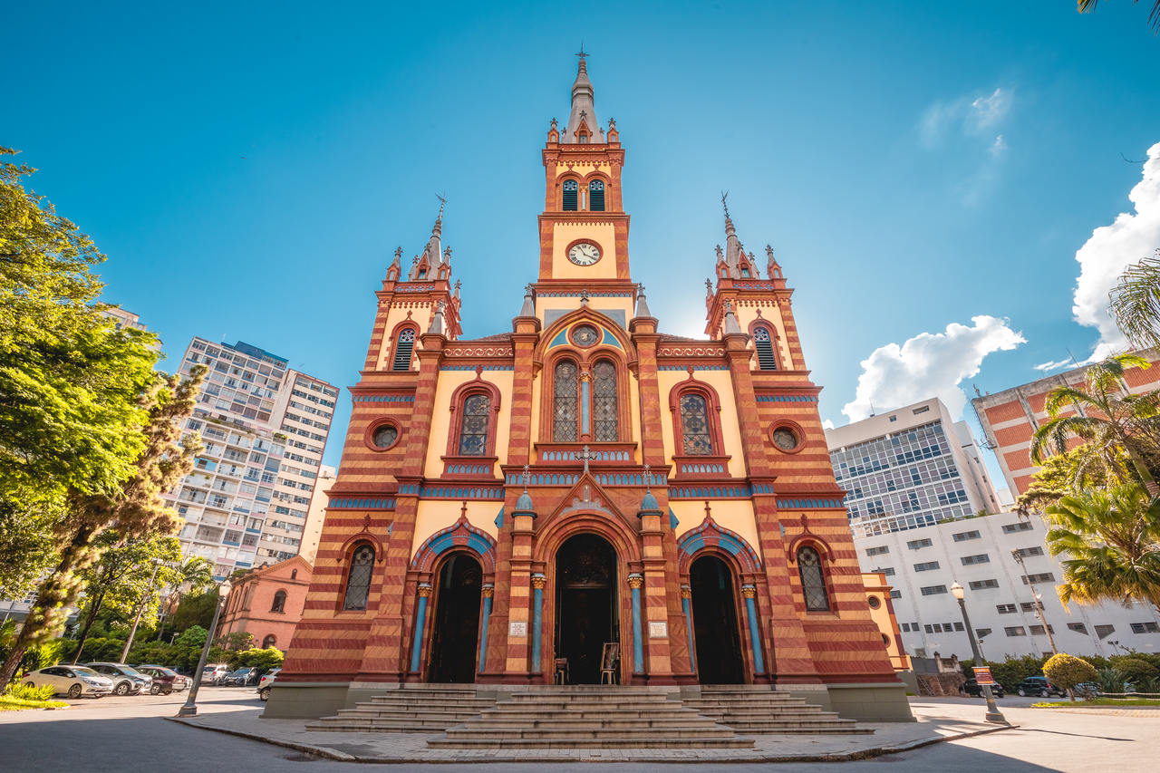 Fachada frontal e simétrica da Igreja de São José em Belo Horizonte. A igreja, de estilo histórico, é de cor vermelha e amarela, com três torres pontiagudas, sendo a central a mais alta e com um relógio. Uma escadaria leva à entrada principal. Ao lado, há prédios altos e árvores, sob um céu azul.