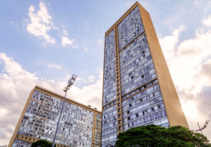 Dois edifícios altos de arquitetura modernista, com fachada envidraçada e laterais em concreto bege. O prédio da direita é mais alto e domina a cena, enquanto o da esquerda aparece parcialmente. Ao fundo, o céu está claro com algumas nuvens, e na parte inferior há árvores e um poste de iluminação urbana.