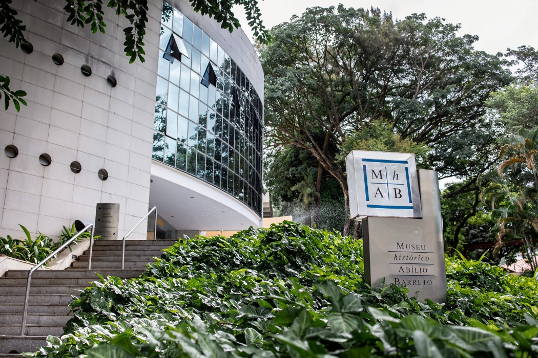 Rafael Costa A entrada moderna do Museu Histórico Abílio Barreto. Em primeiro plano, há um canteiro com plantas verdes e a placa do museu, que exibe o nome e a sigla MHAB. O prédio curvo tem a fachada de concreto cinza e uma grande parede de vidro, que reflete as árvores. Uma escadaria de cimento leva à entrada do edifício.