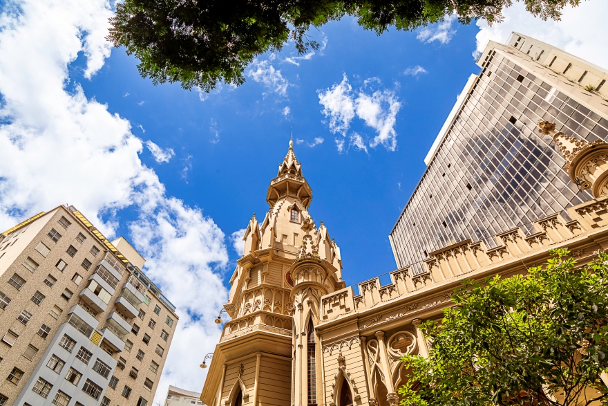 Pablo Gomide/Quarteto Filmes Edifício histórico de estilo gótico, com torre central pontiaguda e ornamentos detalhados, visto de baixo contra um céu azul com nuvens. Ao redor, há prédios modernos e árvores na parte superior e inferior da imagem.