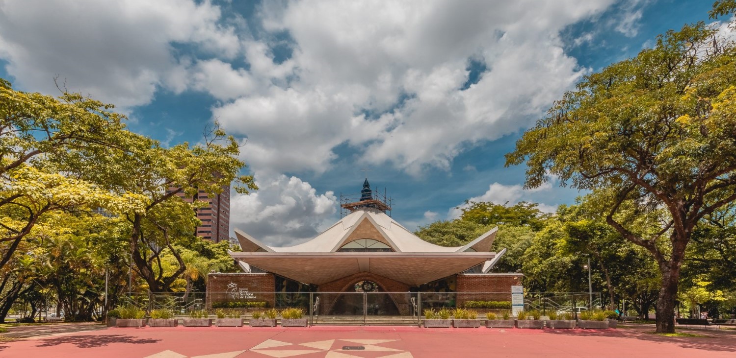 Vista frontal de igreja com telhado branco geométrico em abas inclinadas. No topo, torre azul com cruz. A fachada combina tijolos aparentes e vitrais triangulares. Em frente, canteiros baixos com plantas e piso em vermelho e bege. Árvores grandes cercam o espaço.