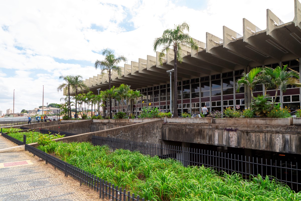 Pablo Gomide/Quarteto Filmes Vista lateral externa do edifício da rodoviária. A marquise de concreto, com seu design de vigas em série, cobre a fachada de vidro. Em frente ao prédio, há um jardim em desnível com palmeiras e vegetação baixa, separado da calçada por uma mureta de concreto e uma grade de metal preta.
